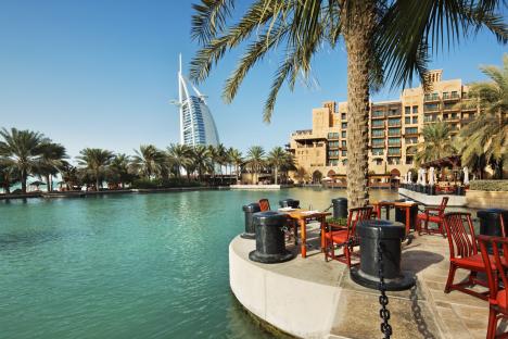 Outdoor restaurant tables and chairs by a canal with the Burj Al Arab hotel in the background.