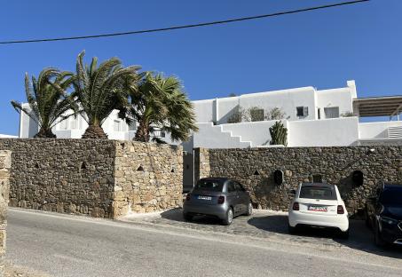 White building with stone wall and palm trees, two cars parked in front.