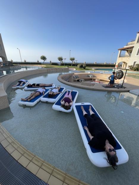 People floating on mats in a pool, with a person playing a gong in the background.