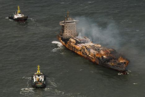 Aerial view of a burning cargo ship being towed by two tugboats.