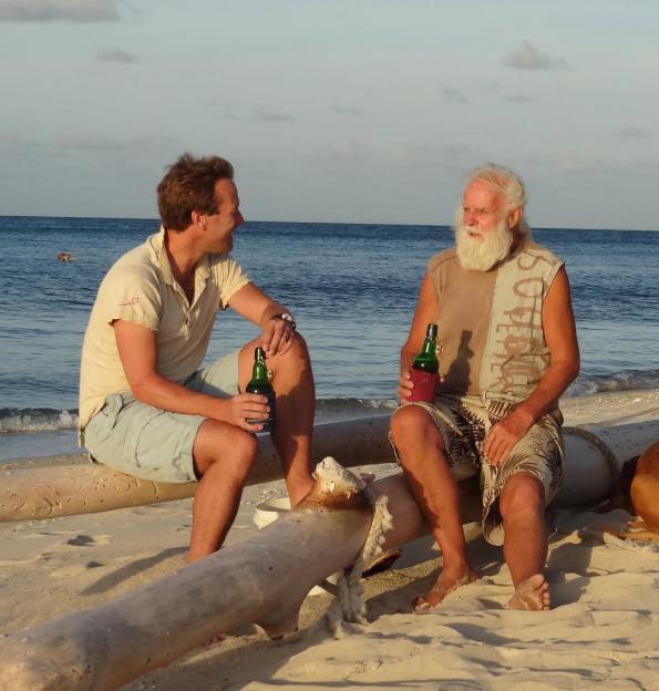 Two men sitting on a log on a beach, each holding a bottle.