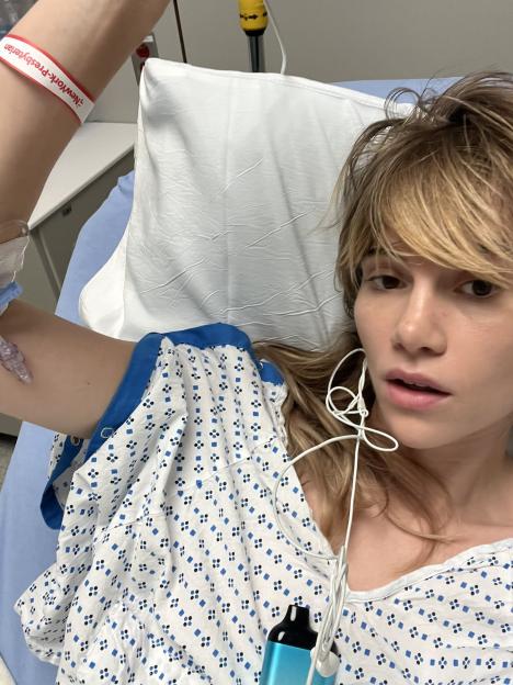 Suki Waterhouse in a hospital gown in a hospital bed.