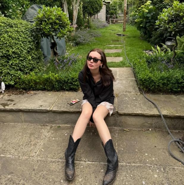 Young woman wearing cowboy boots sitting on stone steps in a garden.