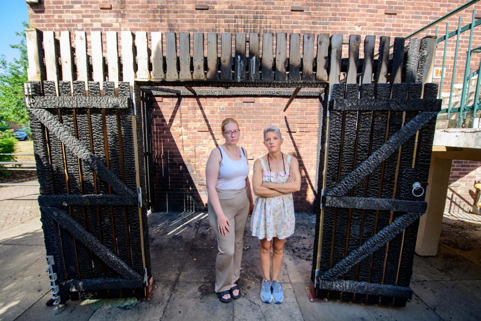Two women stand in front of a fire-damaged gate.