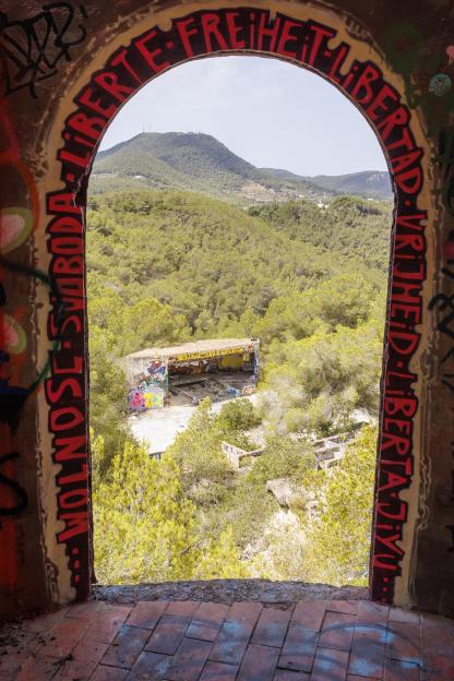 View through graffiti-covered archway of overgrown ruins and distant hills.