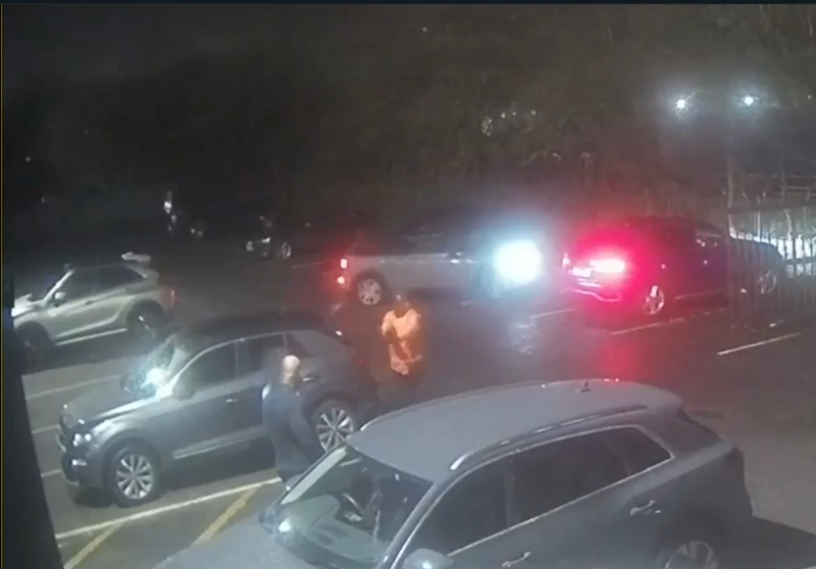Nighttime security camera footage of a parking lot with several cars and two people.