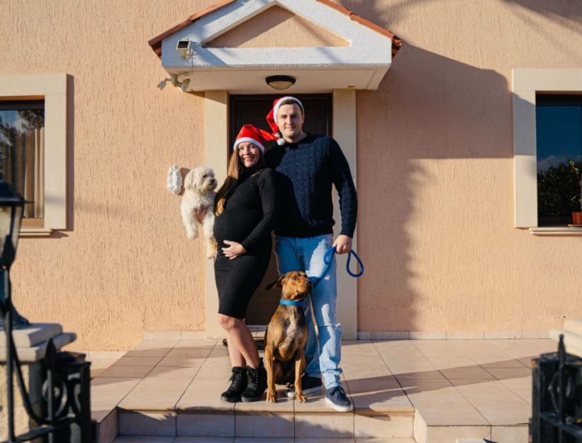 Pregnant woman and man in Santa hats stand outside their home with two dogs.