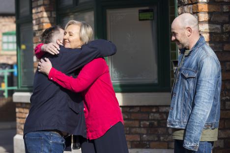 Still image from Coronation Street showing Eileen Grimshaw hugging Steve McDonald and Tim Metcalfe, with George Shuttleworth looking on.