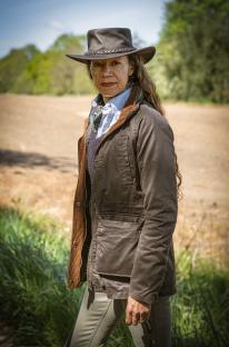 A woman in a brown jacket and hat stands in a field.