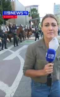 Reporter reporting live from a street with mounted police in the background.