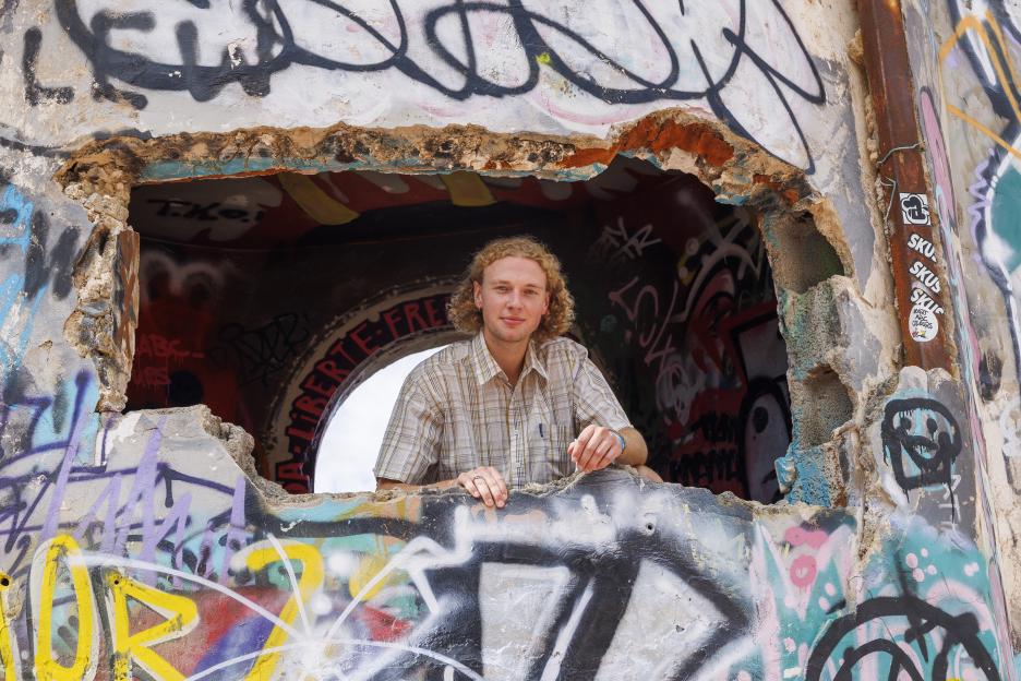 A young man looks out from a hole in a graffiti-covered wall.