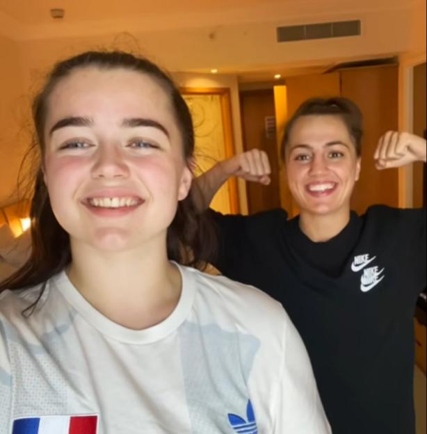 Two women flexing their biceps, one wearing a white shirt with a French flag.