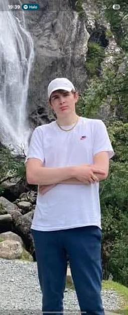 A young man in a white shirt and blue pants stands in front of a waterfall.
