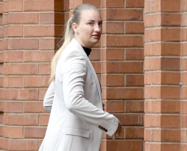 Woman in a light beige blazer walking past a brick wall.