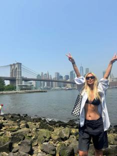 Woman making peace sign in front of the Brooklyn Bridge and New York City skyline.