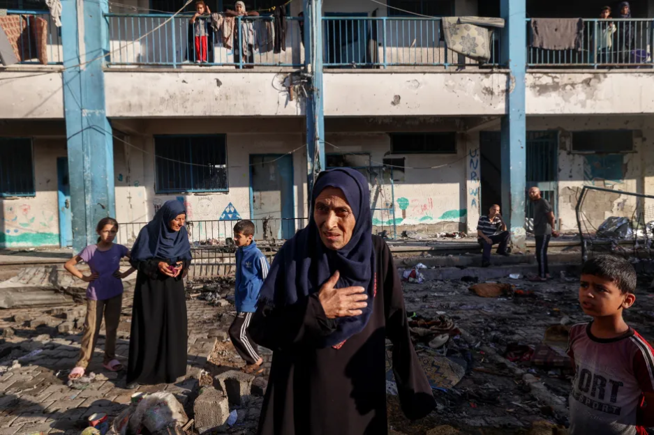 A woman stands amidst rubble, other people visible in the background.
