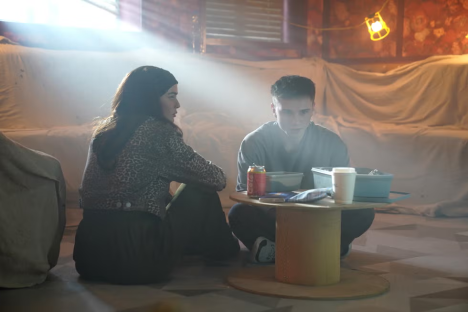 Two young adults sit on the floor, talking.