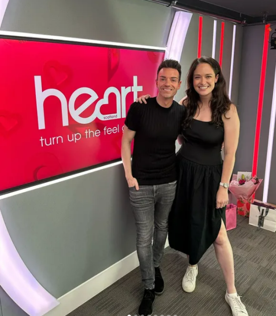 Two people posing for a photo in front of a Heart Scotland radio station backdrop.