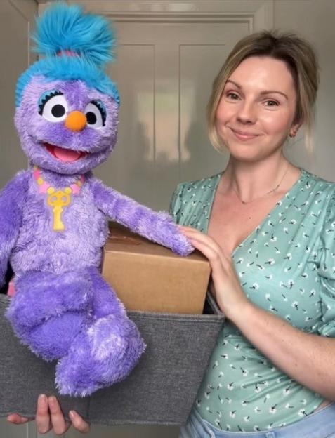 Woman holding a purple puppet and boxes in a storage bin.
