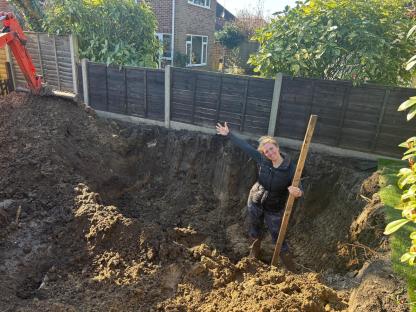 Woman stands in a large hole dug in her backyard, arms outstretched.