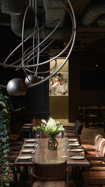 Interior view of INCH Restaurant, showing a long wooden table set for a meal, with leather chairs and a modern chandelier.