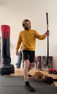 Man holding a samurai sword, skateboard, and cat.