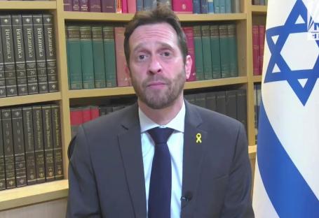 Man in suit speaks in front of bookshelf and Israeli flag.