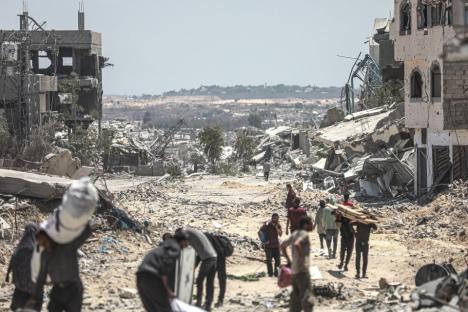 People carrying belongings walk through rubble-strewn streets amidst destroyed buildings.