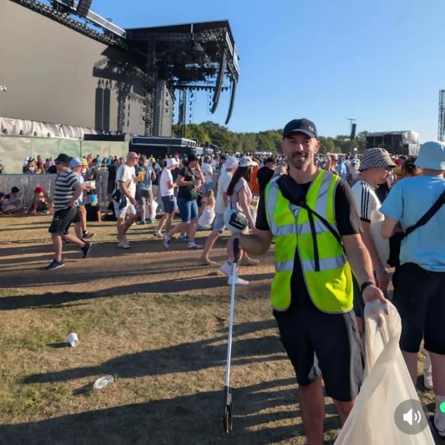 Man in high-visibility vest and cap holding a litter picker and trash bag, with two men walking in the background.