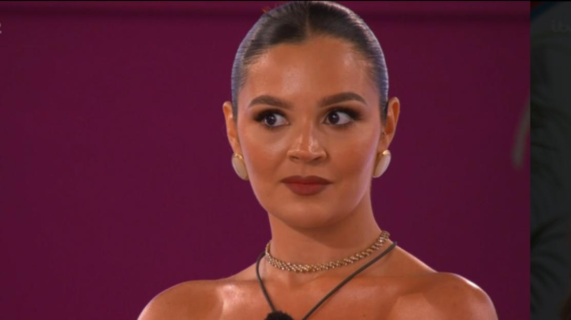Screenshot of a woman with dark hair pulled back, wearing a gold necklace and earrings, against a purple background.