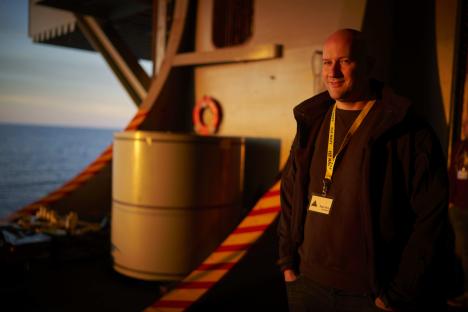Man in dark jacket with lanyard, standing on a ship's deck at sunset.