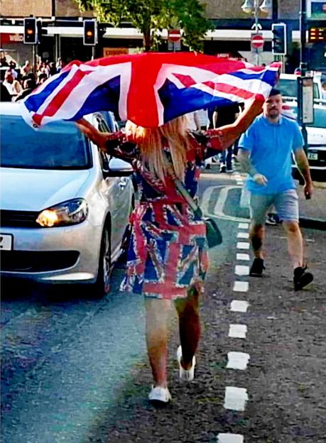 Woman in Union Jack dress walking down a street, holding a Union Jack flag above her head.