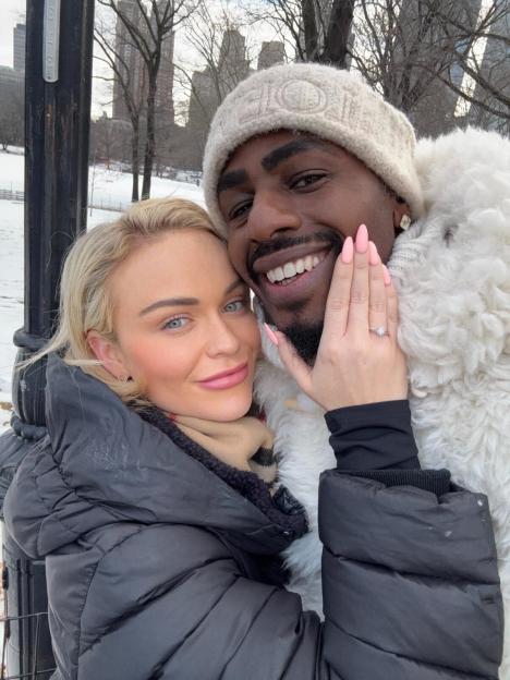 Couple embracing in snowy Central Park; woman showing off engagement ring.