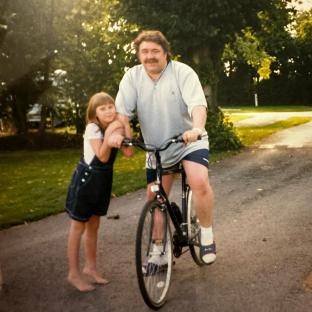 Photo of a father and daughter; the father is on a bicycle, and the daughter is standing next to him.