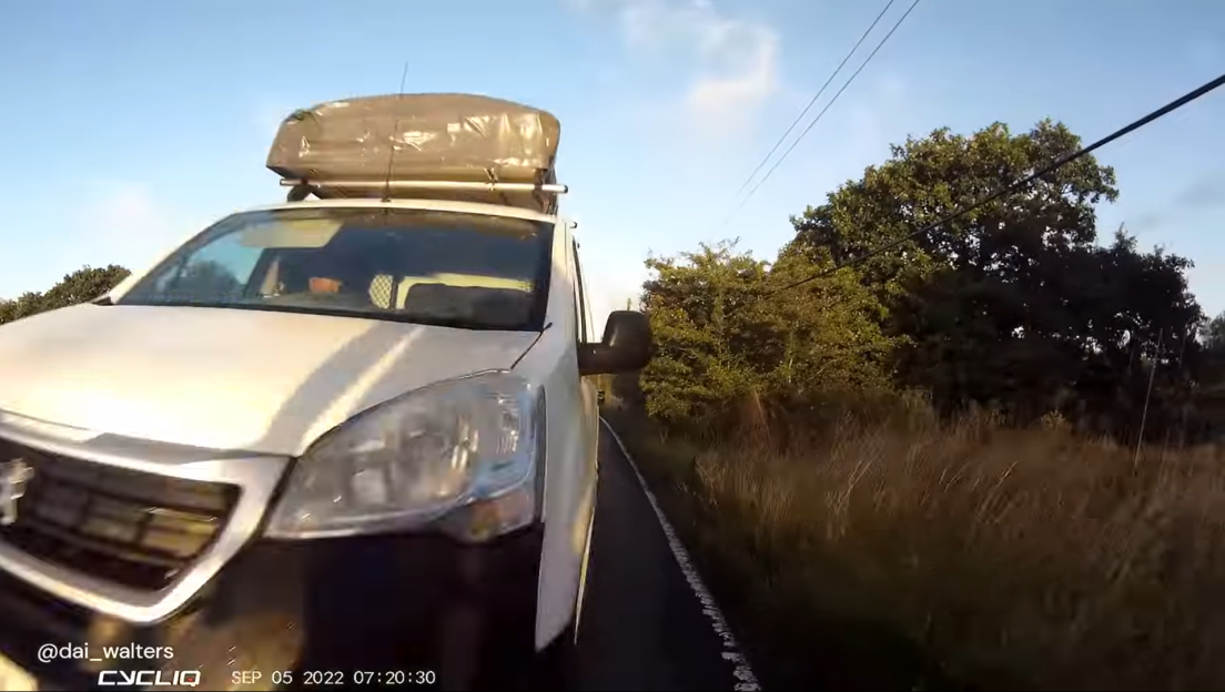 Screenshot of a white van with a roof rack driving down a country road.