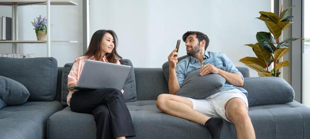 A young couple relaxes on a couch, one using a laptop and the other a phone.