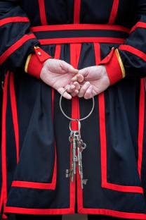 A person in a black and red uniform holds a large set of keys behind their back.