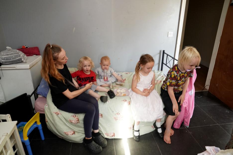 A mother and her four young children sit on a makeshift bed in a cluttered room.