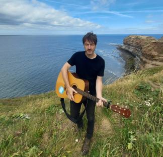 Man playing acoustic guitar on a cliff overlooking the ocean.