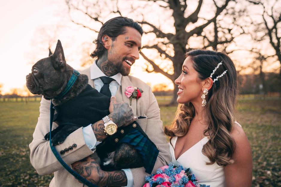 A newlywed couple looks at each other while holding their French bulldog.