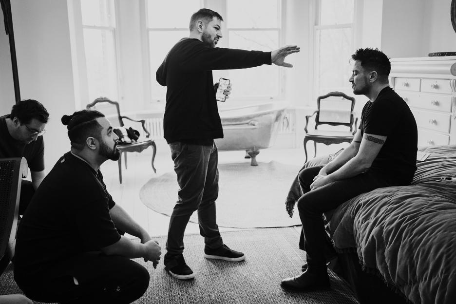 Black and white photo of a photoshoot; a man sits on a bed while another man directs him.