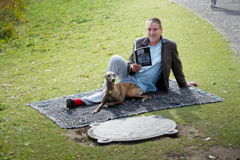 Man reading a book about manhole covers with his dog beside him on a blanket.