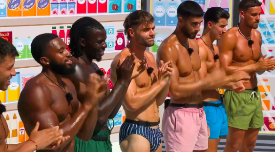 Seven shirtless men clapping in front of a colorful backdrop.