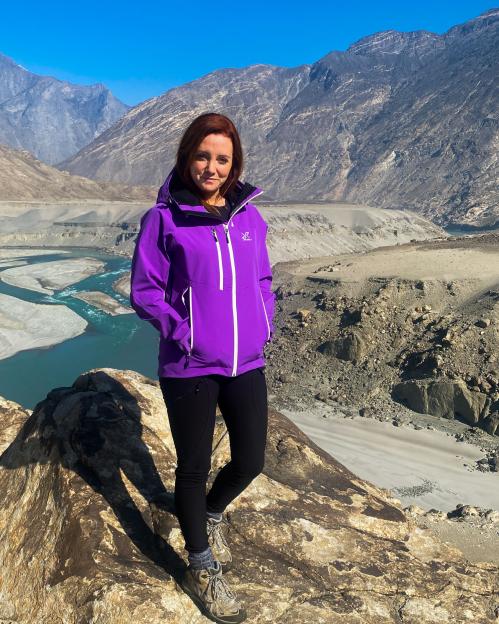 Woman in purple jacket standing on a rock overlooking a mountain river valley.