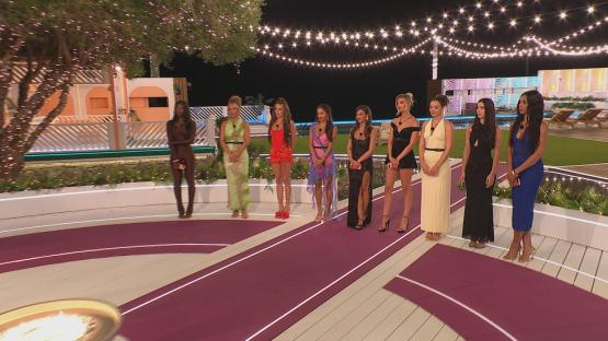 Eight women in evening dresses stand on a red carpet at a nighttime event.