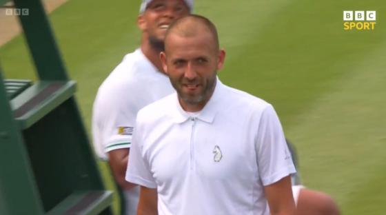 A man in a white polo shirt smiles on a tennis court.