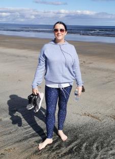 Woman standing barefoot on a beach, holding running shoes and a water bottle.
