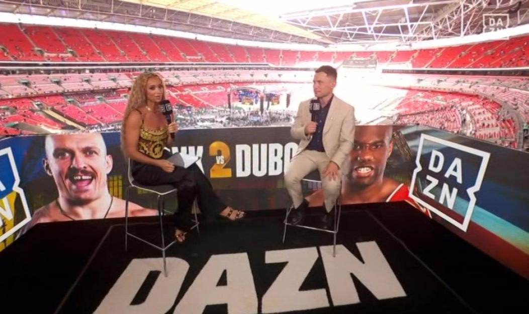 Two sportscasters sitting in front of a large screen showing a boxing match in a stadium.