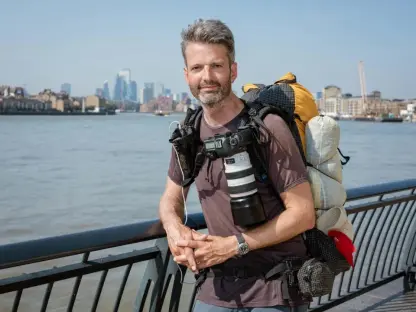 Man with a large backpack and camera, standing by a river with a city skyline in the background.