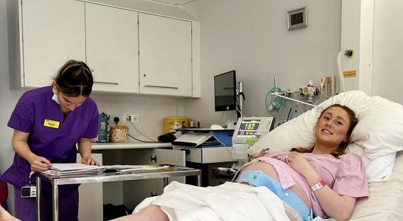 A pregnant woman in a hospital bed, monitored by medical equipment, with a midwife reviewing paperwork.